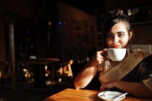 Elegant brunette south asian indian girl in saree posed indoor cafe and drinking tea. photo