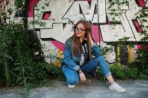 Stylish casual hipster girl in jeans wear and glasses against large graffiti wall. photo