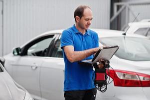 Car repair and maintenance theme. Electric mechanic in uniform working in auto service, making car diagnostics using obd device with laptop. photo