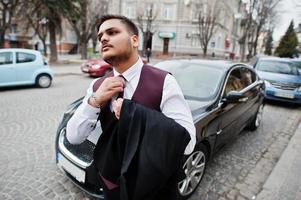 Stylish indian businessman in formal wear tied his tie and standing against black business car on street of city. photo
