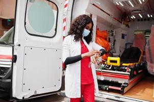 African american female paramedic in face protective medical mask standing in front of ambulance car. photo