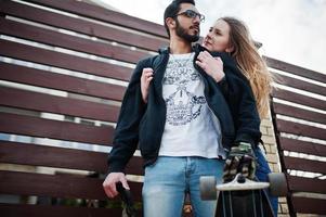 Cool multiracial couple hugs together with longboard against wooden fence. photo