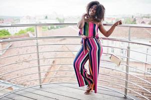 Fashionable african american woman in pink striped jumpsuit, with handbag posed in the french balcony. photo