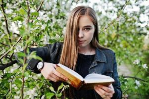 Young brunette girl at jeans against spring blossom tree read the book. photo