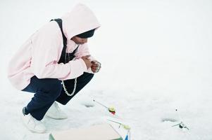 African american fisherman with fishing rod sitting on frozen sea. Winter fishing. photo