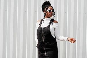 African woman in denim overalls skirt, white sunglasses posed against white steel wall. photo