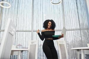 Happy and exults winner fashionable young beautiful african american business woman with afro hairstyle wear in elegant black, stand with laptop at hands. photo