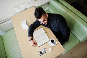 el hombre árabe usa una chaqueta de jeans negros y anteojos sentados en un café, lee un libro y bebe café. chico modelo árabe elegante y de moda. foto