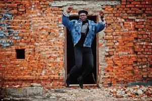 African american man in jeans jacket, beret and eyeglasses against brick wall jump at abandoned roof. photo