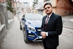 Rich indian businessman in formal wear standing against business suv car. photo