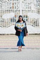 African american dark skinned plus size model posed in a blue shiny dress and black fur coat with bouquet of flowers against large white gates of mansion. photo