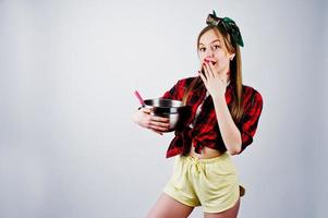 Young funny housewife in checkered shirt and yellow shorts pin up style with saucepan and kitchen spoon isolated on white background. photo