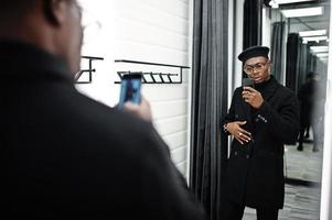 Stylish casual african american man at black beret and overcoat at fitting room clothes store, making photo at mirror on his phone camera.