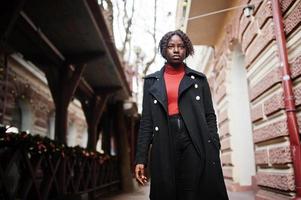 Portrait of a curly haired african woman wearing fashionable black coat and red turtleneck walking outdoor. photo