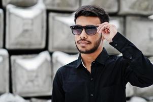 Casual young indian man in black shirt and sunglasses posed against stone blocks. photo