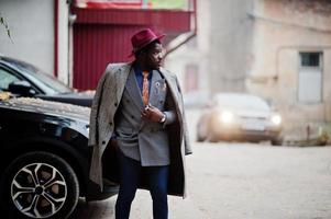 Stylish African American man model in gray coat, jacket tie and red hat against black suv car. photo