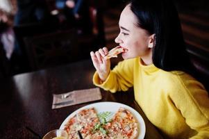 chica morena divertida en suéter amarillo comiendo pizza en el restaurante. foto