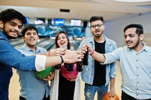 Group of five south asian peoples having rest and fun at bowling club. Clinking cold soda drinks from glass bottles and bowling balls at hands. photo