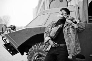 African american man in jeans jacket, beret and eyeglasses, smoking cigar and posed against btr military armored vehicle. photo