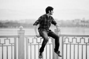 Young indian student man at checkered shirt and jeans sitting on handrails against lake. photo