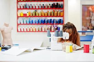 Young african dressmaker woman sews clothes on sewing machine at tailor office. Black seamstress women. photo