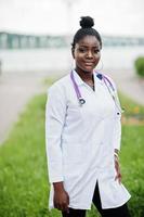 joven doctora afroamericana en bata blanca con un estetoscopio posado al aire libre. foto