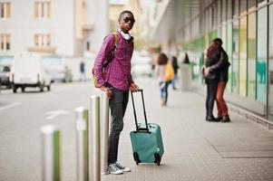 African american man in checkered shirt, sunglasses and earphones with suitcase and backpack. Black man traveler. photo