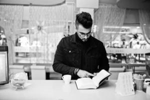 el hombre árabe usa una chaqueta negra de jeans y anteojos en un café bebe café en el bar con un libro. chico modelo árabe elegante y de moda. foto