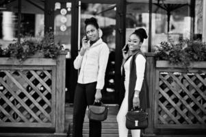 Two stylish african american girls friends with handbags posed outdoor of steets city and speaking on mobile phone. photo