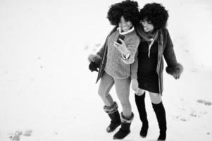 Dos mujeres afroamericanas de pelo rizado se visten con abrigo de piel de oveja y guantes posados en el día de invierno y haciendo selfie por teléfono. foto