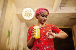 Stylish african woman in red shirt and hat posed indoor cafe and drinking pineapple lemonade. photo