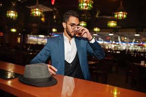 apuesto hombre árabe bien vestido con vaso de whisky y cigarro posado en el pub. foto