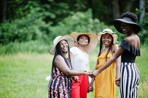 Group of four gorgeous african american womans wear summer hat holding hands and praying at green grass in park. photo