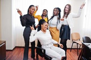 cinco mujeres de negocios multirraciales de pie en la oficina y ruedan a la mujer en la silla. grupo diverso de empleadas en ropa formal divirtiéndose. foto