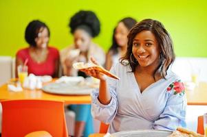 African woman with pizza sitting at restaurant against dark skinned girls. photo