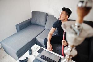 Handsome and fashionable indian man in black sitting at room, smoking hookah, working on laptop. photo