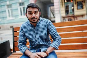 Stylish indian beard model man in casual clothes posed outdoor, sitting on bench at street of India and looking at his watches. photo