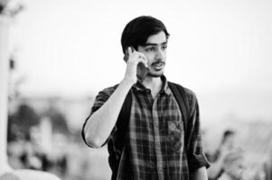 Young indian student man at checkered shirt and jeans with backpack speaking on mobile phone. photo