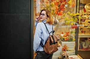 Cheerful young beautiful redhaired woman in glasses, warm blue wool sweater with backpack posed outdoor against autumn leaves tree decorations on store. photo