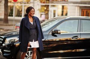 éxito elegante mujer afroamericana en abrigo contra coche suv de negocios negro.