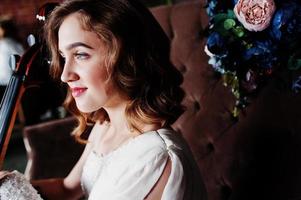 Pretty young gilrl musician in white dress with double bass sitting on brown vintage sofa. photo