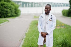 joven médico afroamericano de bata blanca con un estetoscopio posado al aire libre. foto