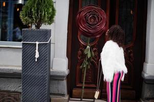 Fashionable african american woman in pink striped jumpsuit with fluffy faux fur coat and handbag posed at street against large rose. photo