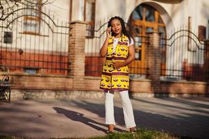 Stylish african american women in yellow jacket posed on street at sunny day with mobile phone at hand. photo