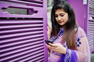 Indian hindu girl at traditional violet saree posed at street against purple windows with mobile phone at hands. photo