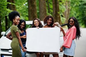 Group of five happy african american girls posed against car and hold empty white blank banner. Free space for your text. photo