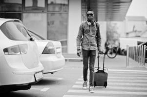 African american man in checkered shirt, sunglasses and earphones with suitcase and backpack. Black man traveler walking on crosswalk. photo