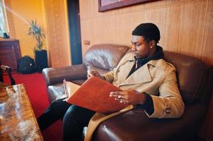 Handsome african american man posing  inside room with sunlight shadows in black hat and beige coat. photo