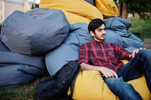 joven estudiante indio con camisa a cuadros y jeans sentado y relajado en almohadas al aire libre. foto