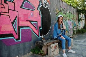 Stylish casual hipster girl in cap and jeans wear listening music from headphones of mobile phone against large graffiti wall with bomb. photo